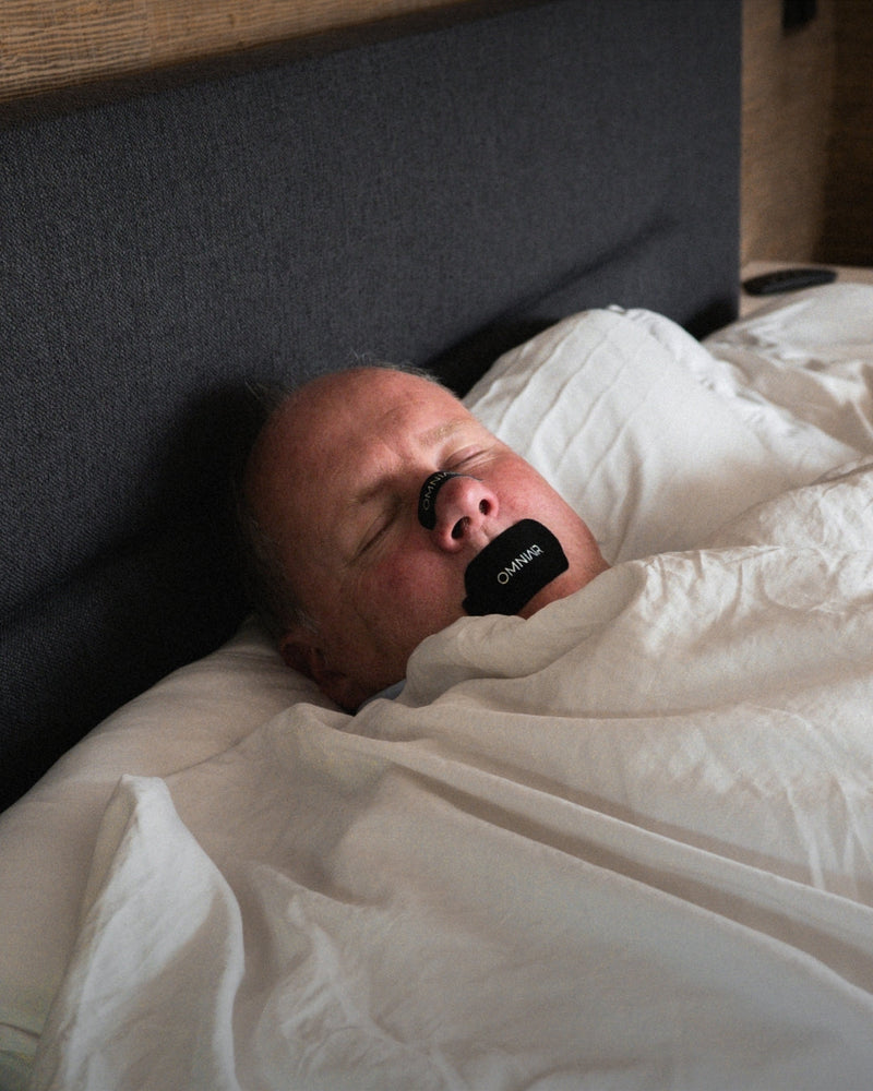 Man sleeping in bed with black OMNIAIR mouth tape across his mouth, breathing through the nose.